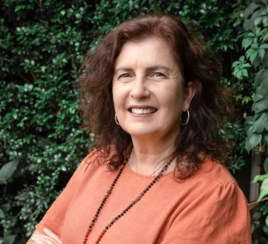 Head shot of Travel Wiz, Kerri Seccombe, standing outside in the shade of green garden shrubs brimming with a big welcoming smile ready to take in all the detail to planning your next holiday.