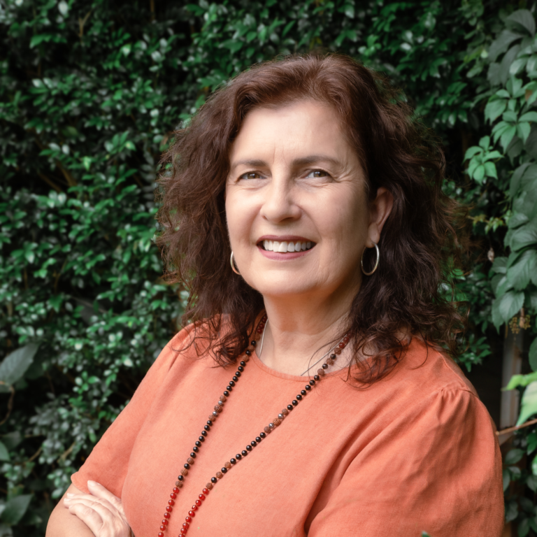 Head shot of Travel Wiz, Kerri Seccombe, standing outside in the shade of green garden shrubs brimming with a big welcoming smile ready to take in all the detail to planning your next holiday.