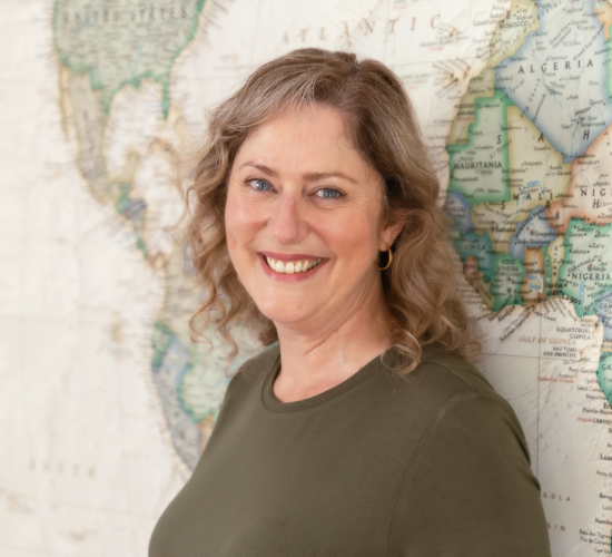 Headshot of a smiling Marketing Coordinator for Icon Adventures, standing confidently in front of a world map. Laurene is looking directly at the camera, conveying professionalism and warmth.
